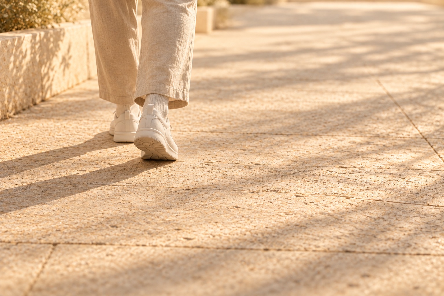 Lower legs in sand-coloured trousers and cream trainers walking along a quiet sunlit stone path.